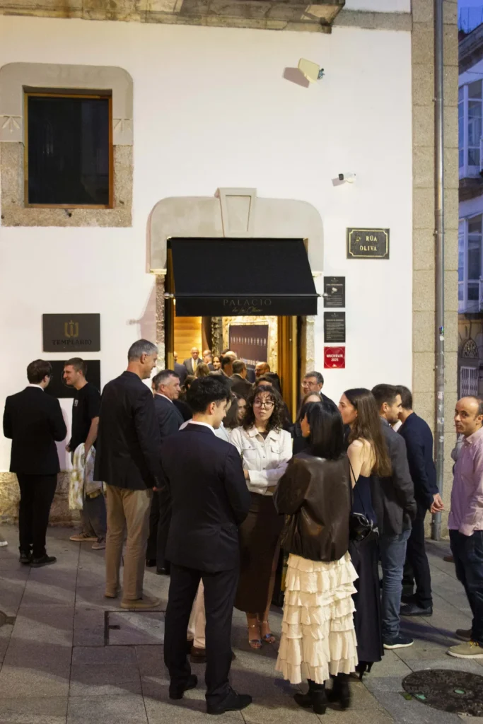 Convidados conversan en pequenos grupos á entrada iluminada dun edificio branco ao anoitecer